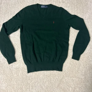 Mörkgrön tröja från Polo Ralph Lauren - Säljer en stilren mörkgrön tröja från Polo Ralph Lauren med v-ringning och broderad logotyp på bröstet. Tröjan har långa ärmar och ribbade muddar vid ärmslut och nederkant. Perfekt för en klassisk look.
