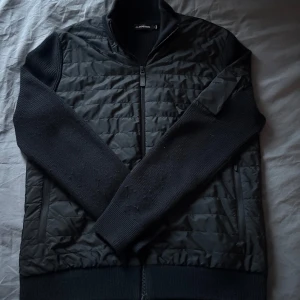 Svart Cardigan från J.Lindeberg - Snygg svart cardigan från J.Lindeberg med quiltad framsida och dragkedja. Ärmarna och ryggen är i ett ribbat material som ger en stilren look. Använd i endast en säsong så är i väldigt bra skick. Nypriset låg på drygt 3200kr