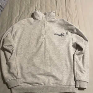 Boohoo 1/4 zip up  - Säljer en stilren grå tröja från Boohoo med halv dragkedja och broderad text på bröstet. Tröjan har långa ärmar och ribbade muddar, perfekt för en avslappnad look. Köpt i Malmö på en Pop-up store, sparsamt använd