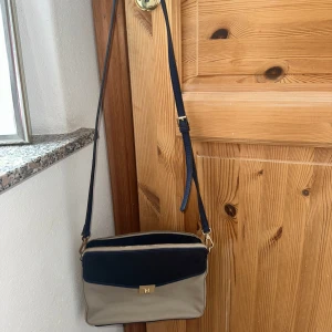 Beige och marinblå axelväska - Snygg axelväska i beige och marinblått med gulddetaljer. Väskan har en praktisk dragkedja och en justerbar axelrem. 