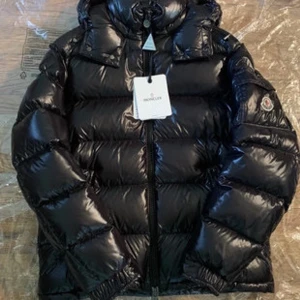 Svart dunjacka från Moncler - Snygg och varm svart dunjacka från Moncler med hög krage och dragkedja. Perfekt för kalla dagar med sin pufferdesign och blanka yta. Jackan har långa ärmar och en klassisk Moncler-logga på ärmen.