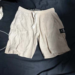 Beige shorts från Calvin Klein - Snygga beige shorts från Calvin Klein med snörning i midjan och ett diskret märke på sidan. Perfekta för en avslappnad stil.