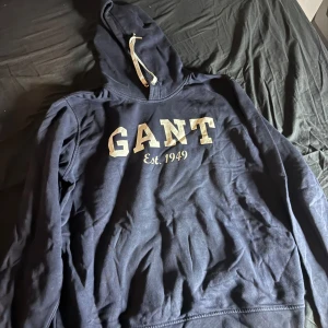 Mörkblå hoodie från GANT - Säljer en snygg mörkblå hoodie från GANT med texten 'GANT Est. 1949' i vitt på framsidan. Den har en klassisk huva med dragsko och långa ärmar. Perfekt för en avslappnad stil.