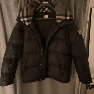 Vinter jacka  - Stilren och varm Burberry-dunjacka i svart med klassisk rutmönstrad insida av kragen. Jackan är i storlek XL men fungerar som L och passar perfekt för kalla dagar. Den är mångsidig och kan även förvandlas till en väst tack vare avtagbara ärmar – perfekt för mildare väder. Jackan har två praktiska dragkedjefickor och snygga detaljer, inklusive märkets logga 'Burberry London England' på ärmen. Jackan är i bra skick och håller hög kvalitet, typiskt för Burberrys produkter. Behöver kemtvättas