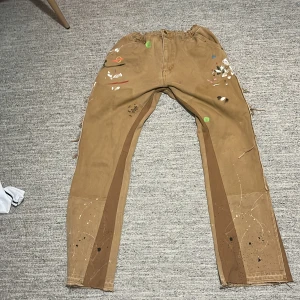 Bruna arbetsbyxor med färgstänk - Säljer ett par bruna arbetsbyxor med unika färgstänk i olika färger. Byxorna har flera praktiska fickor och en elastisk midja för extra komfort. Perfekta för kreativa projekt eller arbete där stil möter funktion.