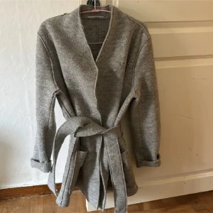 Grå kappa från Filippa K - Säljer en stilren och mysig grå ullkappa från Filippa K i storlek small. Kappan har en elegant omlott-design med ett bälte i midjan för en snygg passform.  En knapp borta kan lätt fixas❤️ Köpt den förra året ca 4000kr