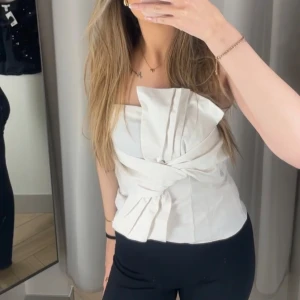 Vit axelbandslös topp - Snygg vit axelbandslös topp med volangdetaljer framtill. Perfekt för en stilren och elegant look. Toppen har en unik design med knytning i midjan som ger en smickrande passform.