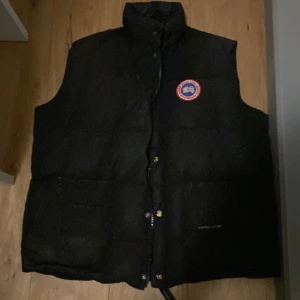 Svart dunväst från Canada Goose - Säljer en svart dunväst från Canada Goose med ett broderat märke på bröstet. Västen har knappar och är perfekt för kyligare väder.