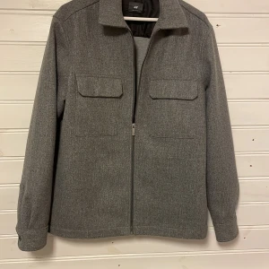 Grå overshirt från H&M - Snygg grå overshirt från H&M med dragkedja och två bröstfickor med lock. Perfekt för lager-på-lager-stil under höst och vår. Den har en klassisk krage och långa ärmar.