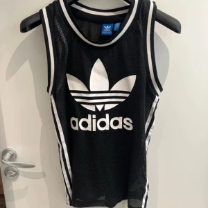 Svart ärmlös topp från Adidas - Snygg svart ärmlös topp från Adidas med det klassiska logotyptrycket i vitt. Tillverkad i ett mesh-liknande material som ger en sportig känsla. Perfekt för träning eller en avslappnad stil.