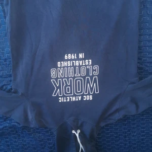 Mörkblå hoodie från SOC - Säljer en mörkblå hoodie från SOC med texten 'WORK CLOTHING' och 'ESTABLISHED IN 1989' tryckt på framsidan. Tröjan har en klassisk design med dragsko i huvan och långa ärmar. Perfekt för en avslappnad stil.