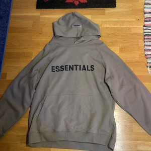 Beige Essentials hoodie från Fear of God - Säljer en beige Essentials hoodie från Fear of God. Den har en stor ficka framtill och texten 'ESSENTIALS' tryckt på bröstet. Perfekt för en avslappnad stil. Tröjan är helt oanvänd och helt ny med lapp kvar.