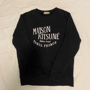 Svart sweatshirt från Maison Kitsuné - Snygg svart sweatshirt från Maison Kitsuné med vit text på framsidan. Perfekt för en stilren och avslappnad look. Tröjan har rund hals och långa ärmar. Är i storlek XL men passar bättre som L eller stor M