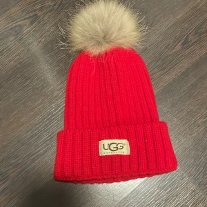 Röd stickad mössa från UGG - Snygg röd stickad mössa från UGG med en fluffig pälsboll på toppen. Mössan har en ribbad design och ett uppvikt kant med UGG-logotypen framtill. Perfekt för att hålla värmen under kalla dagar. storlek Small. Är ej använd kommer som ny. Var inte riktigt min typ av mössa till mig. Men den är jättefin och varm nu till vintern🫶🏼💕skriv vid funderingar o frågar:) 
