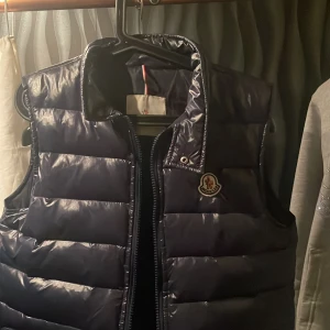 väst Moncler - Snygg marinblå dunväst från Moncler med dragkedja och hög krage. Perfekt för kyligare dagar och ger en stilren look. Västen fick jag av mina föräldrar för många år sedan och den är från Carl of Carl har dock inte andvändt den så jätte mycket då den varit för stor.
