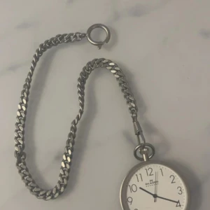 Silverfärgad fickur med kedja - Elegant silverfärgat fickur från Mr. Beaumont med en klassisk vit urtavla och tydliga svarta siffror. Uret har en matchande kedja för att fästa i fickan eller västen. Perfekt för en stilfull och tidlös look.