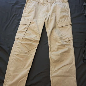 Beige cargobyxor från Dressmann - Säljer ett par beige cargobyxor från Dressmann med flera praktiska fickor och knappstängning. Byxorna har en normal passform och är perfekta för en avslappnad stil.