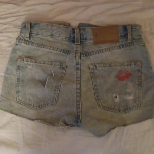 Blå jeansshorts - Säljer ett par blå jeansshorts från newyotker storlek XXS. Shortsen har en sliten look med revor och en cool detalj med ett rött läppstiftsmärke på ena bakfickan. Perfekta för en avslappnad stil! Högmidja