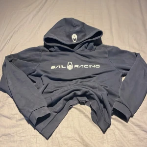 Mörkblå hoodie från Sail Racing - Säljer en mörkblå hoodie från Sail Racing med vit logga på bröstet. Tröjan har en klassisk design med huva och långa ärmar. Perfekt för en avslappnad stil.