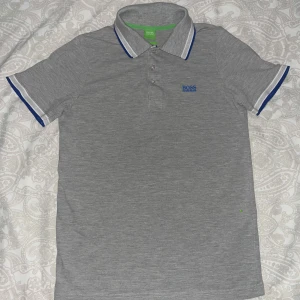 Grå pikétröja från Boss - Snygg grå pikétröja från Boss med blå och vita detaljer på kragen, använd några gånger men är som ny och den har inga skador säljer den för 600 pris kan diskuteras.