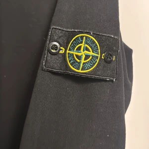 Svart tröja från Stone Island - Snygg svart tröja från Stone Island med det ikoniska märket på ärmen. Tröjan har en rund hals och långa ärmar, perfekt för en stilren look.