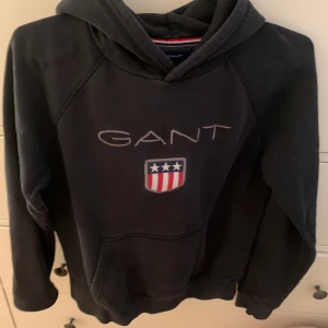 Svart hoodie från GANT - Säljer en svart hoodie från GANT med deras logga och ett emblem på bröstet. Tröjan har en klassisk design med huva och långa ärmar. Perfekt för en avslappnad stil.