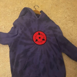 Lila hoodie med anime-tryck - Cool lila hoodie med Naruto-tryck på baksidan och ett rött emblem på framsidan. Perfekt för fans av Naruto! Hoodien har en stor ficka framtill och en bekväm huva.