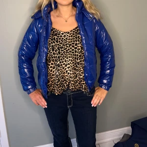 Blå glansig pufferjacka - Snygg blå pufferjacka med glansigt material och dragkedja framtill. Jackan har en hög krage och långa ärmar, perfekt för kyligare dagar. Den har en modern och trendig look som passar bra till jeans. ( den är liten i storleken, snarare S!)