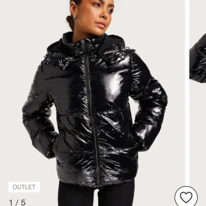 Svart glansig pufferjacka - Snygg svart glansig pufferjacka med dragkedja och huva. Perfekt för kyliga dagar med sin vadderade design. 