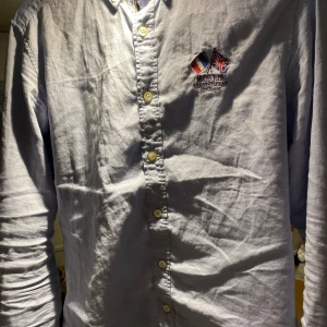 Ljusblå skjorta från Morris - Snygg ljusblå skjorta från Morris med broderade flaggor på bröstet. Skjortan har knappar framtill och är tillverkad i 100% bomull, vilket gör den bekväm och andningsbar. Perfekt för sommaren inget fel på den den är storlek S men passar mig som har M 