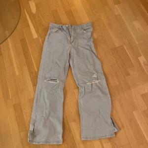 Ljusa jeansbyxor från H&M - Säljer ett par ljusa jeansbyxor från H&M med hög midja och justerbar midja. Byxorna har slitna detaljer på knäna och en bred passform. Perfekta för en avslappnad stil.