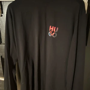 Svart långärmad tröja från Hugo - Säljer en svart långärmad tröja från Hugo med färgglad logotyp på bröstet och ett coolt tryck på ryggen. Perfekt för en stilren och modern look.