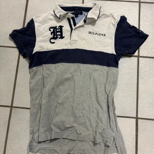 Grå och vit pikétröja från Hilfiger - Snygg pikétröja från Hilfiger i grått och vitt med mörkblå detaljer. Tröjan har korta ärmar och en klassisk krage med knappar. Perfekt för en avslappnad stil.