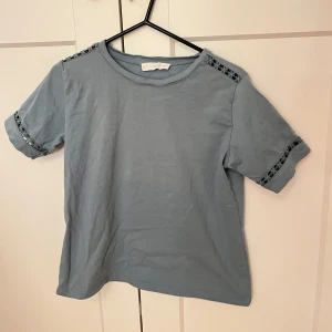 Blå t-shirt med spetsdetaljer - Säljer en ljus-blågrå t-shirt med korta ärmar och fina spetsdetaljer på ärmarna och axlarna. Färgen framgår tydligare på andra bilden!
