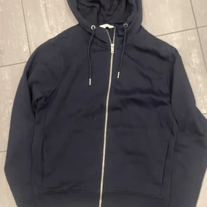 Mörkblå hoodie från Next - Säljer en mörkblå hoodie från Next med dragkedja och justerbar huva. Perfekt för en avslappnad stil. Tillverkad i mjukt material för extra komfort.