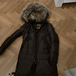 Brun Parajumper Masterpiece Series - Säljer en lång Parajumpers Masterpiece Series med pälsfodrad huva. Jackan har dragkedja och är perfekt för kalla vinterdagar. Den är XS och har praktiska detaljer som gör den både snygg och funktionell.
