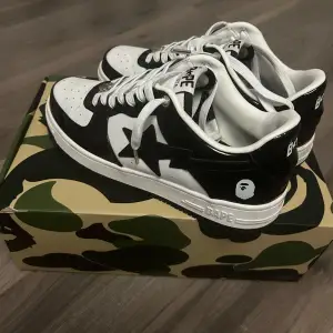 Säljer ett par snygga svartvita sneakers från BAPE. Skorna har en klassisk design med en stjärna på sidan och är i nyskick. De kommer i en cool kamouflagemönstrad låda. Perfekta för att ge din outfit en streetwear-känsla!