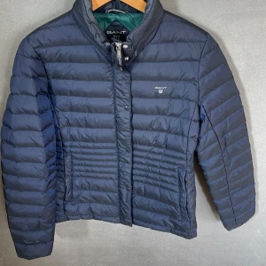 Mörkblå pufferjacka från GANT - Säljer en snygg mörkblå pufferjacka från GANT. Jackan är perfekt för kyliga dagar med sin quiltade design och långa ärmar. Den har en dragkedja framtill och är i nyskick. Passar perfekt för både höst och vinter!