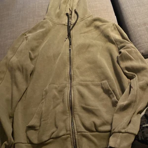 Olivgrön hoodie med dragkedja - Säljer en olivgrön hoodie med dragkedja framtill. Den har en mysig huva och praktiska fickor. Perfekt för en avslappnad stil och passar bra till höst och vår.