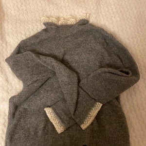 Grå stickad tröja med spetsdetaljer - Säljer en superfin grå stickad tröja med spetsdetaljer vid halsen och ärmsluten. Perfekt för höst och vinter, den är både varm och stilren. Tröjan har långa ärmar och en normal passform. Passar perfekt till både vardag och fest!