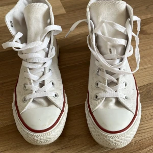 Vita Converse All Star sneakers - Säljer ett par klassiska vita Converse All Star sneakers i canvas. De har en hög modell med snörning och den ikoniska loggan på sidan. Skorna har en röd rand runt sulan och är perfekta för en avslappnad stil. Använda fåtal gånger.