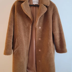 Brun teddyjacka från Stand Studio - Säljer en supermysig brun teddykappa från Stand Studio. Den är perfekt för kalla höst- och vinterdagar. Kappan har långa ärmar och knappar framtill. Insidan är fodrad med ett slätt material som känns skönt mot huden. Den är både stilren och varm, perfekt för att hålla stilen i kylan!