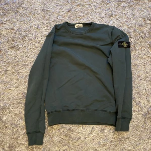 Mörkgrön tröja från Stone Island - Säljer en snygg och stilren mörkgrön tröja från Stone Island. Den har långa ärmar och en klassisk rund halsringning. Tröjan är i bra skick och har det ikoniska Stone Island-märket på ärmen. Perfekt för höst och vinter!