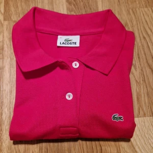 Rosa Lacoste Piké - Denna piké är i färgen rosa och är i storlek 42. Pikén funkar med t shirt under men även utan. Skjortan kommer från Lacoste och är i väldigt bra skick, skjortan har aldrig använts.Pris kan diskuteras.