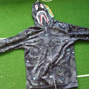 Svart hoodie med hajmotiv - Säljer en cool svart hoodie med ett unikt hajmotiv på huvan. Den har ett camouflagemönster med inslag av blått och vitt, vilket ger den en extra touch. Hoodien har en dragkedja framtill och är perfekt för både vardag och streetwear. Den är i bra skick och redo för nya äventyr!