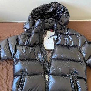 Moncler maya - Säljer en snygg och varm svart pufferjacka från Moncler.Jackan har en glansig finish och är perfekt för kalla vinterdagar. Säljer den på grund av att den är liten för mig. Perfekt för dig som vill hålla stilen även när det är kallt ute! 