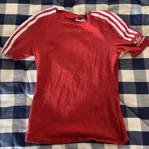 Röd t-shirt från Adidas - Säljer en snygg röd t-shirt från Adidas med de klassiska tre vita ränderna längs ärmarna. T-shirten är kortärmad och gjord i ett mjukt bomullsmaterial. Perfekt för träning eller en avslappnad vardagslook. Den är i bra skick och redo för nya äventyr!