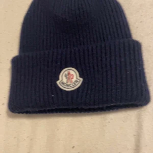 Svart stickad mössa från Moncler - Säljer en snygg svart stickad mössa från Moncler. Den är i nyskick och har en broderad logga framtill. Perfekt för kalla höst- och vinterdagar. Mössan är navy blue och ger en skön passform. Äkta!!!!