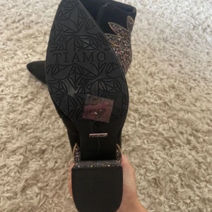 Svarta boots med glittrig klack från Tiamo - Säljer ett par svarta boots från Tiamo med en fantastisk glittrig klack och spetsig tå. De har en dragkedja på sidan för enkel på- och avtagning. Perfekta för att lysa upp vilken outfit som helst!      Helt nya och oanvända 