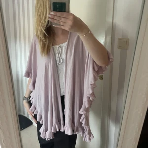 Ljusrosa kofta med volangdetaljer - Säljer en superfin ljusrosa kofta. Den är perfekt för både vår och sommar med sina luftiga volangdetaljer längs ärmarna och nederkanten. Koftan är öppen framtill och har en loose passform, vilket gör den både bekväm och stilren. 